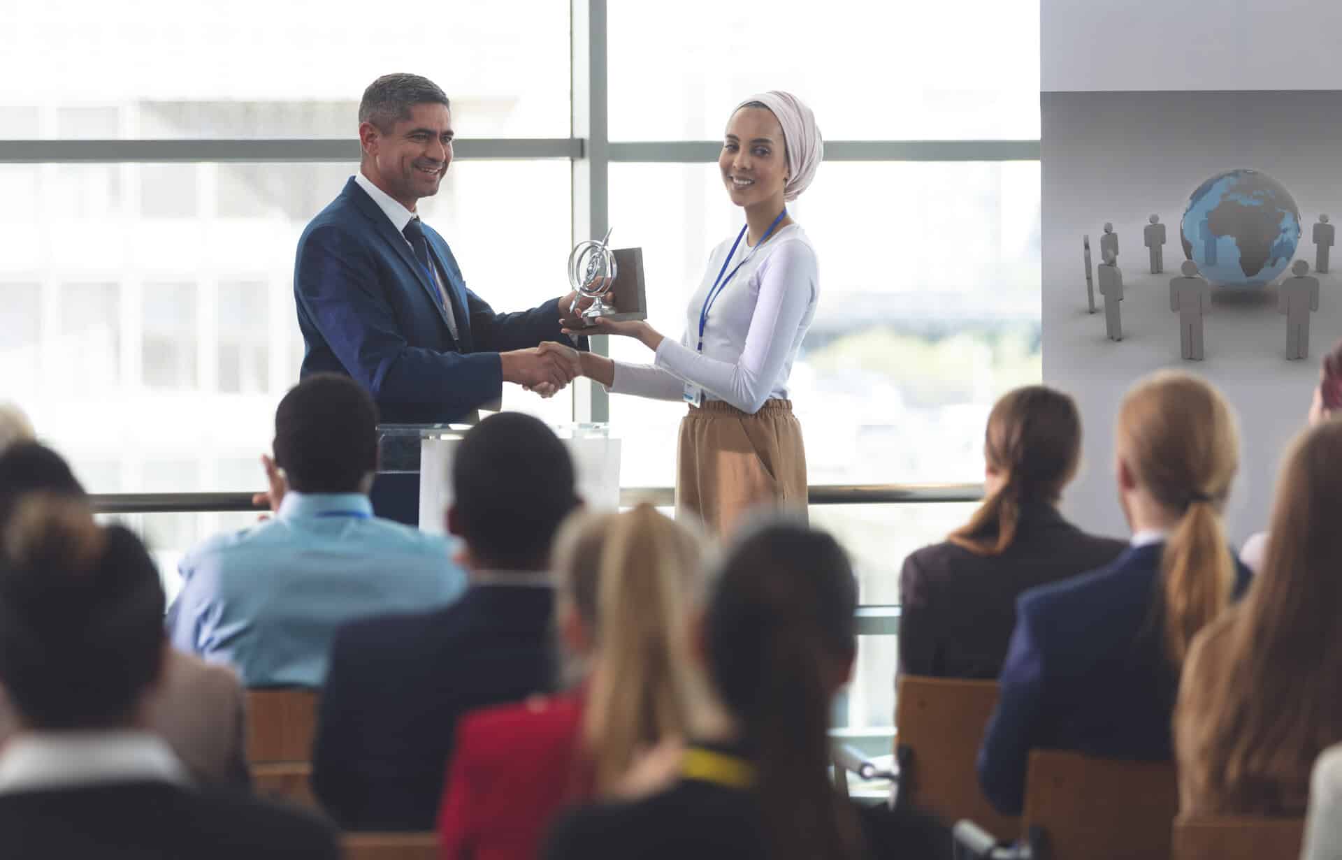 front view of mixed race businesswoman receiving award from mixed race businessman in front of business professionals sitting at a business seminar in office building